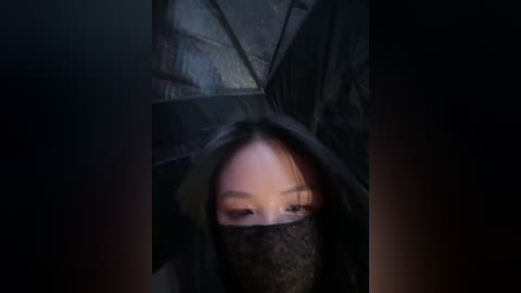 Media: Video of a person with fair skin and short, dark hair partially obscured by a black, textured mask, standing in a dimly lit room with dark, shadowy walls.