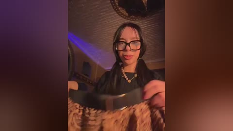 A video of a young Asian woman with short black hair, glasses, and a black jacket, holding a leopard-print purse, in a dimly lit room with a textured ceiling.