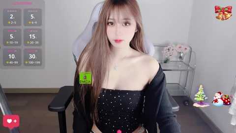 A video of a young Asian woman with long brown hair, fair skin, and wearing a black off-shoulder dress, seated in a gaming chair in a minimalist room with a white wall calendar, a glass shelf with decorative items, and a green overlay.