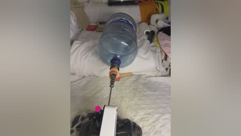 Video of a blue plastic water bottle mounted on a white pillow, connected to a syringe, in a cluttered, messy bedroom with stuffed animals and clothes.