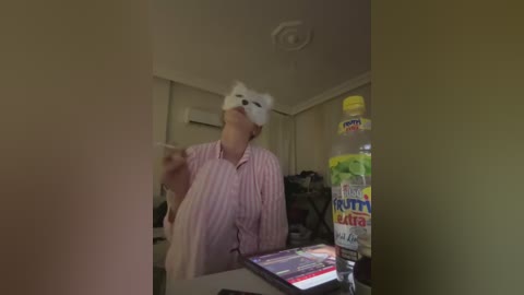 A video of a person wearing a white bear mask and striped pajamas, holding a phone, in a dimly lit room with a large water bottle and a colorful laptop on a desk.