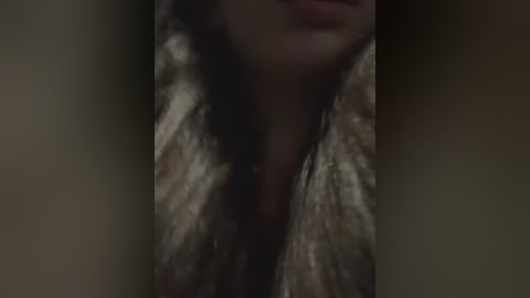 A blurry, dimly-lit close-up video of a woman's face, partially obscured by dark shadows. She has long, dark hair and wears a metallic, glittery jacket. Her lips are slightly parted, hinting at a possible smile.