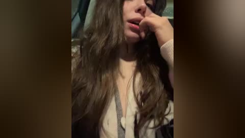 A video of a young woman with long, wavy brown hair, light skin, and a plump lip, seductively biting her finger. She wears a beige cardigan, partially unbuttoned, revealing a black and white striped shirt. The background is blurred, suggesting an indoor setting.