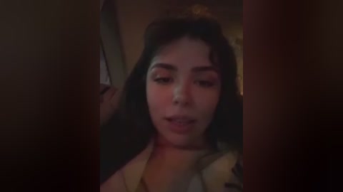 A dimly lit video captures a young woman with dark hair and fair skin, partially obscured by a shadowy figure in the foreground.