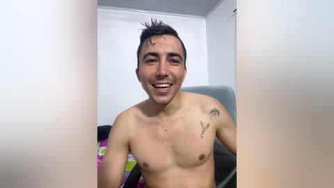 Video of a shirtless, smiling young man with short dark hair, tattoos on his chest, and a light skin tone, sitting on a chair in a minimalist room.