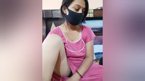 Video of a young woman with medium brown skin, wearing a pink dress, black face mask, and black hair, seated on a bed, blurred background.