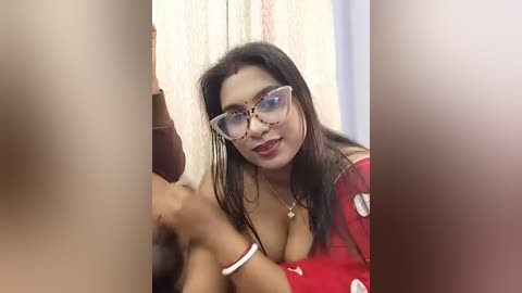 Video of a South Asian woman with long black hair, wearing glasses and a red top, sitting on a bed.