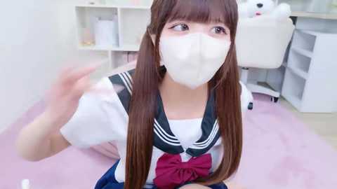 A young woman with long brown hair, wearing a school uniform, white mask, and a pink bow, kneels on a pink carpet in a bright, minimalist room with white furniture and shelves.