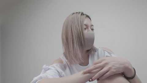 Video of a young Asian woman with short, straight, platinum blonde hair, wearing a white top and gray face mask, sitting against a plain, light gray wall. She has her hands on her chest, appearing calm and contemplative.
