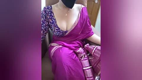 A video of a woman in a bright pink sari with a floral blouse, seated on a brown couch, wearing a black face mask.