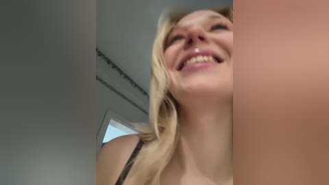A video of a smiling blonde woman with fair skin and long, wavy hair, wearing a black lace bra, captured from a low angle. Background shows a simple, modern room with a door and white walls.
