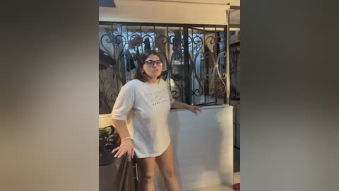 Video of a young woman with short dark hair, wearing glasses and a loose white T-shirt, standing by a wrought-iron gate in a cozy, dimly lit room.