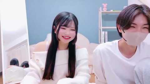 Video of two young Asian women in white lab coats and masks, smiling, in a bright, modern medical office with blue walls and shelves.