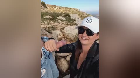 Media: Video of a smiling woman with sunglasses, wearing a white baseball cap and black leather jacket, holding a man's erect penis. Background features rocky terrain and blue sky.