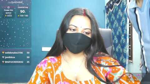 Video of a South Asian woman with long black hair, wearing a black mask, orange floral blouse, and seated in a salon chair. Background features a blue wall, a weather forecast screen, and hanging clothes.