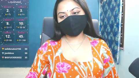 Video of a woman with long dark hair, wearing a black face mask, floral orange top, and a blue wall with a calendar and curtain in the background.