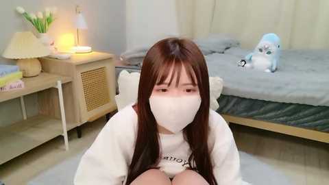 Video of an Asian woman with long brown hair wearing a white mask, white hoodie, and seated on a light-colored rug in a cozy bedroom with a bed, nightstand, and stuffed toys.