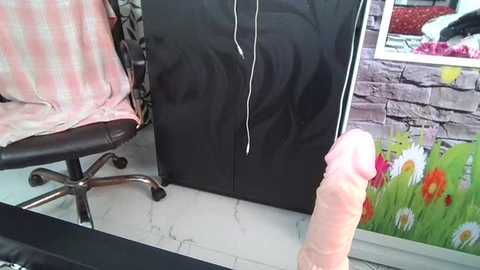 Video of a person holding a large, realistic-looking dildo, standing next to a black gym bag and a pink plaid chair, with a colorful floral mural in the background.