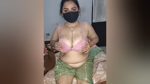 Video of a South Asian woman with medium skin tone, dark hair, wearing a pink lace bra, green saree, and black face mask, sitting on a bed with floral sheets in a dimly lit room.