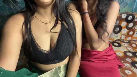 Video of two women, one wearing a black lace bra and another a red saree, on a patterned couch, capturing a casual, intimate moment.