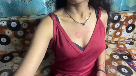 Video of an Asian woman with light skin, dark hair, wearing a red tank top and black bra, seated on a couch with a patterned cushion. She has a bruise on her left shoulder.