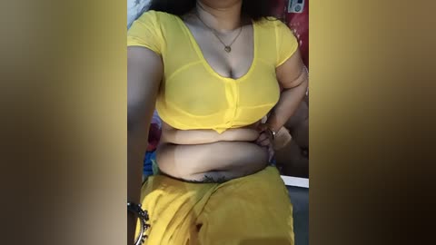 A video of a South Asian woman with medium skin tone, wearing a sheer yellow blouse and yellow skirt. She has a curvy figure, a small tattoo on her lower abdomen, and a gold necklace. Background shows part of a red wall.