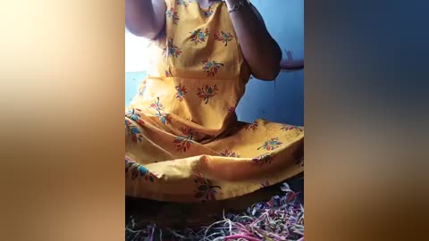 A video of a person in a yellow dress with floral patterns, seated on a colorful rug. The background is a plain, light blue wall. The image is framed by a soft, blurry gradient.