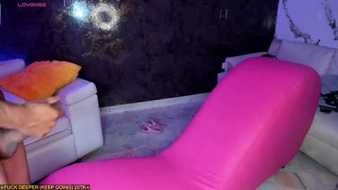 A video of a pink inflatable lounge chair and a white couch with a fluffy orange pillow in a dimly lit room with dark walls.