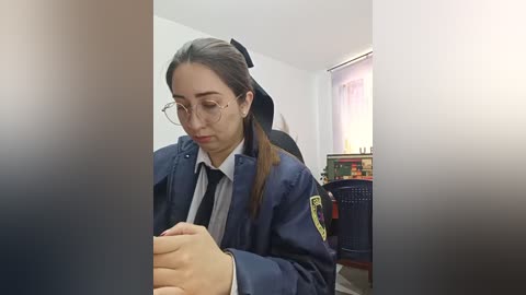 Video of a young woman with light skin and long brown hair in glasses, wearing a navy school uniform, looking down at a smartphone, seated in a room with a window and bookshelf.