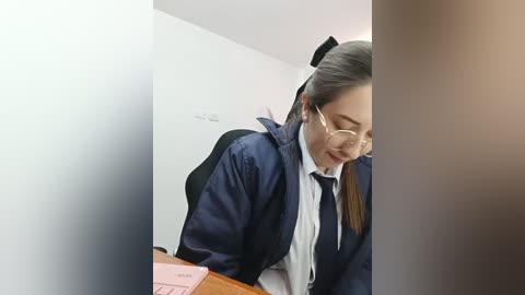 Video of an Asian woman with light skin and long black hair tied back, wearing a navy blue jacket over a white shirt and black tie. She's seated at a desk in a dimly lit room with white walls.