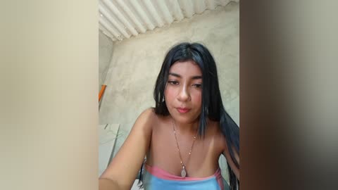 Video of a young woman with long black hair, wearing a strapless top, standing in a rustic, corrugated metal-roofed room with a concrete wall.