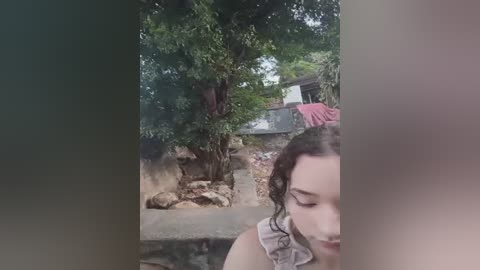 A video depicts a young woman with curly dark hair and pale skin, wearing a sleeveless top. She is partially visible in the foreground, while a rustic, greenery-filled backyard with a stone wall and a red clothed structure is seen in the background.