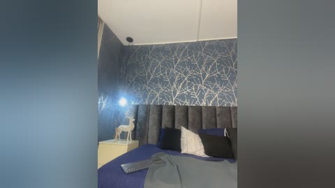 Video of a modern bedroom with a blue-and-white tree-patterned wallpaper, a gray upholstered headboard, a white deer figurine on a nightstand, and a remote control on the bed.