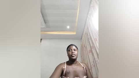 Video of a dark-skinned, medium-built woman with a round face, wearing a light pink bra. She stands in a modern room with white walls, a ceiling light, and a partially visible patterned curtain.