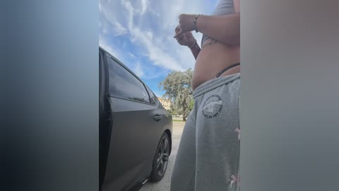 Video of a woman in grey sweatpants, standing next to a black car in a suburban neighborhood. She has a toned midriff, holding a camera, under a partly cloudy sky.
