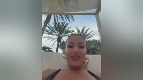 Media: Video of a smiling woman with light skin, wearing a black bikini top, reclining in a sunbed under a white canopy, with a clear blue sky and palm trees in the background.