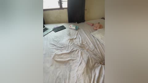 A video shows a messy bedroom with a laptop, a keyboard, and a box on a bed, a baby lying on the bed, and a window with a dark curtain.