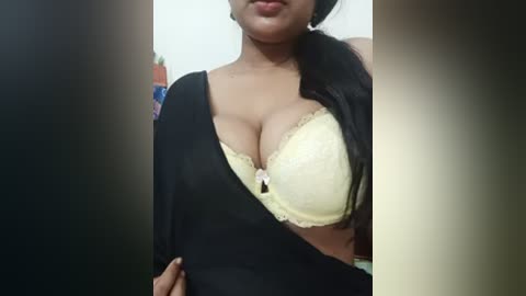 Video of a South Asian woman with medium brown skin, wearing a black sari draped over a yellow lace bra, showcasing her ample cleavage. Background shows a blurred indoor setting with a white wall.