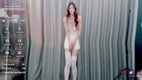 Video of a slim Asian woman with long, wavy red hair, wearing a white, strapless dress and white thigh-high stockings. She stands against a backdrop of vertical, light-colored curtains.