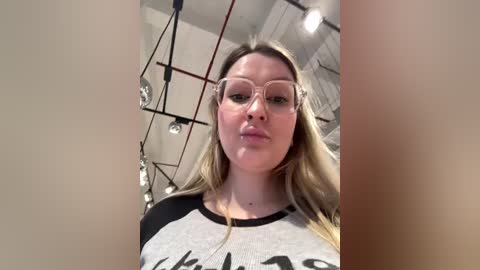 Video of a blonde woman with fair skin, wearing clear glasses and a black-and-white graphic tee, taken from a low angle in a studio with industrial lights and a grid ceiling.