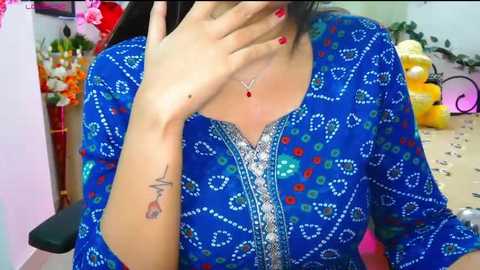 Media: Video of a woman with light skin, dark hair, wearing a vibrant blue, floral-patterned shirt with intricate embroidery, and red nail polish. Background includes a pink floral arrangement, yellow balloons, and a black chair.