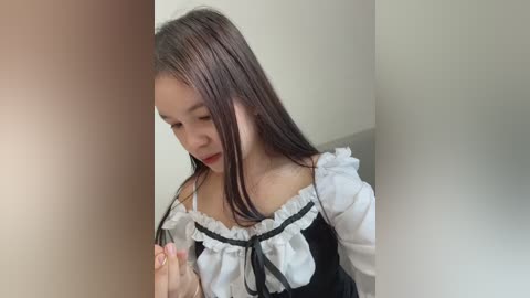 Video of a young Asian woman with long, straight, dark hair, wearing a traditional black and white French maid outfit, looking down and holding a small object in her hands.