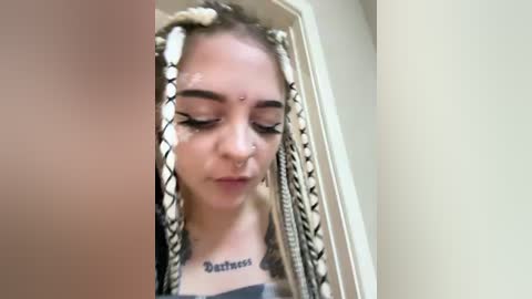 Video of a young white woman with light skin, long braids, and a nose ring, looking down, in a beige room with a white door.