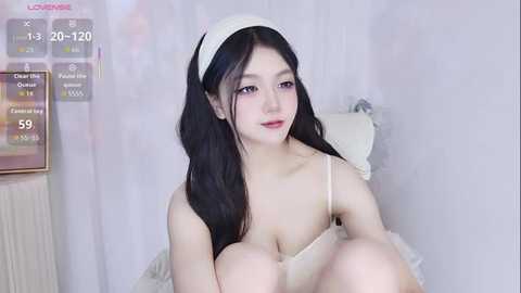Video of a young Asian woman with long black hair, wearing a white headband and a revealing white lingerie set, sitting on a white bed with a pale pink background.