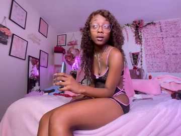 Video of a slim, dark-skinned woman with long curly hair, wearing glasses, pink lingerie, and holding a sex toy, sitting on a bed in a pink-themed room with framed certificates and a brick wall.