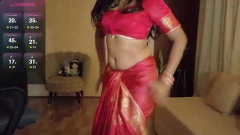 Video of a woman wearing a red sari with gold embroidery, standing in a living room with a wooden floor and beige furniture, including a plush chair and sofa.