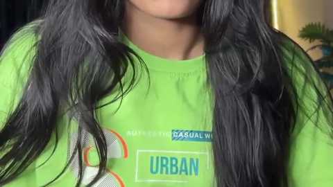 Video of a person with long, straight black hair, wearing a bright green t-shirt with the word \"URBAN\" in blue. The background is blurred, showing a room with a lamp.