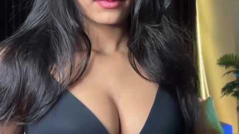 Video of a woman with long, black, wavy hair, wearing a black, low-cut top, showcasing her ample cleavage. Background features a blurred, indoor setting with a plant.