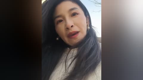 Video of a young woman with long, straight black hair, wearing a white top, looking up with a neutral expression, taken outdoors with a bright, clear sky in the background.