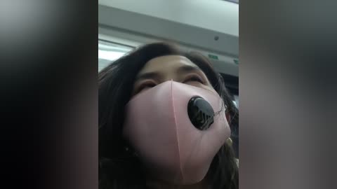 A video of an Asian woman wearing a pink surgical mask with a black eye patch over her right eye, taken from a low angle, emphasizing her closed eyes and mask. The background is a blurred indoor setting with a ceiling light.